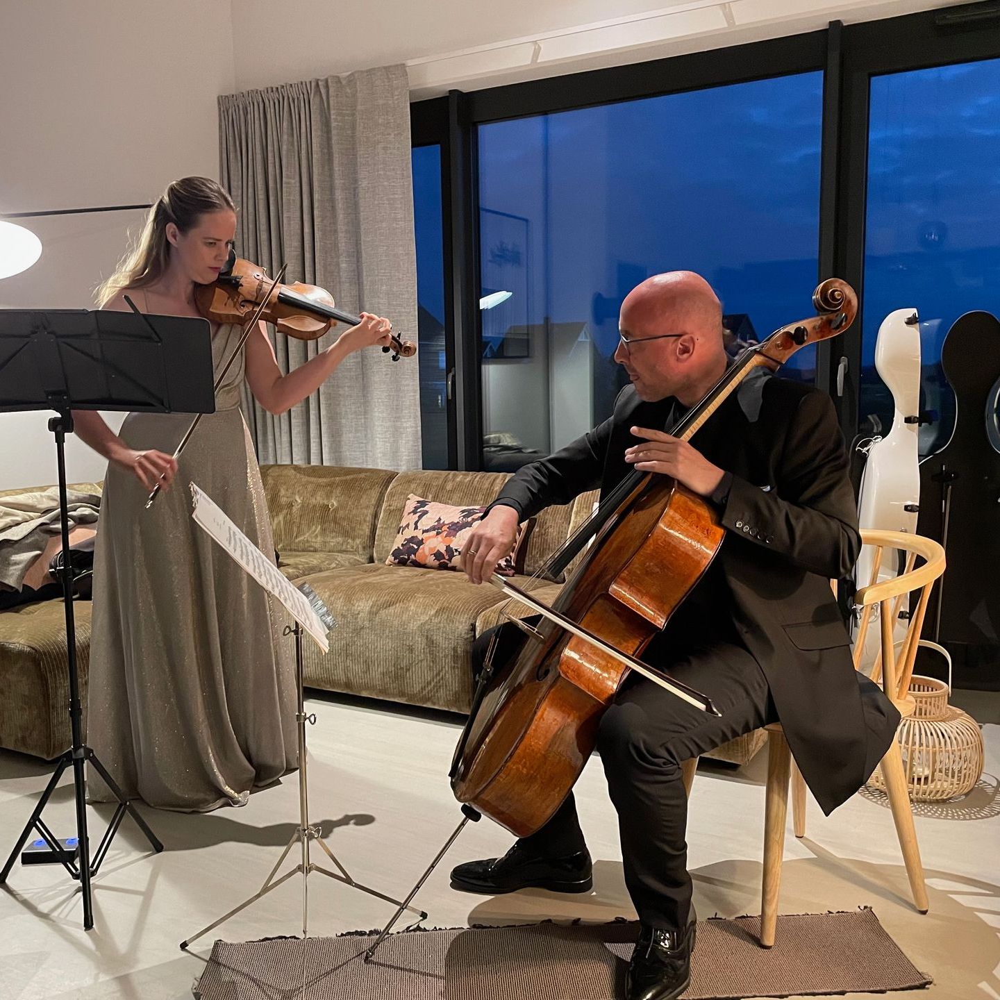 In einem Wohnzimmer spielt ein Mann Cello, während eine Frau Geige spielt.