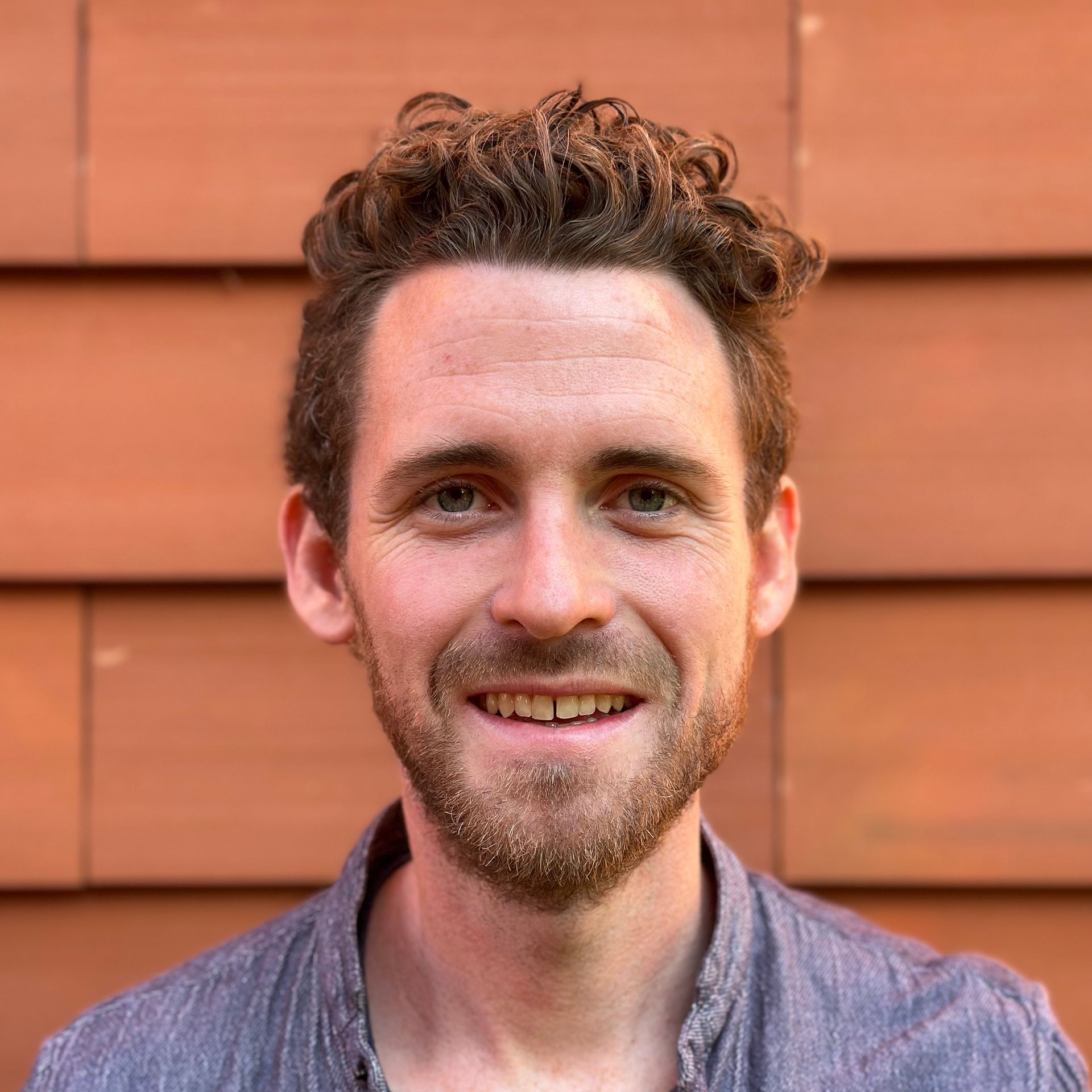 A man with curly hair and a beard is smiling in front of a red wall.