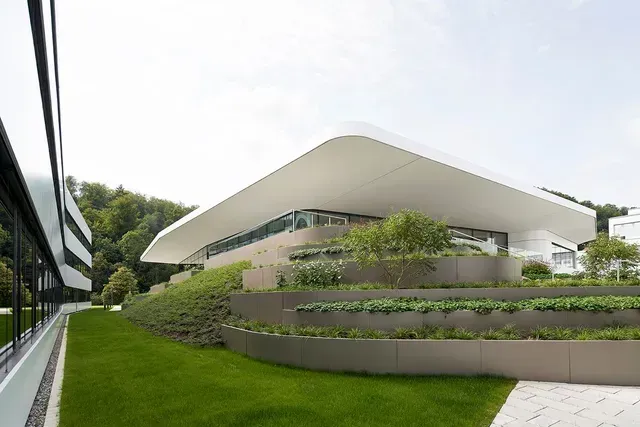 Ein großes weißes Gebäude mit einem geschwungenen Dach ist von Gras und Bäumen umgeben.