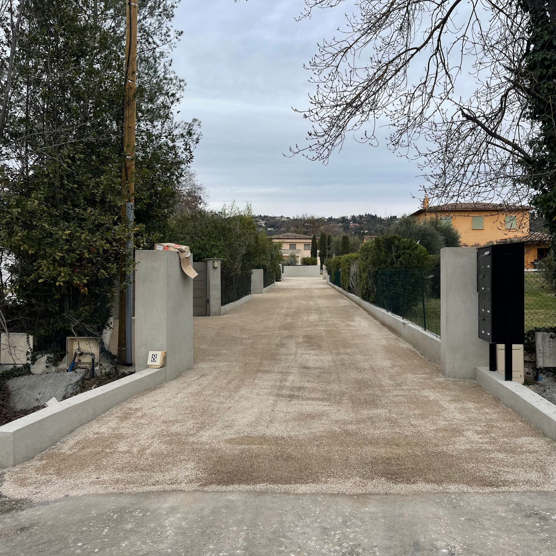 Allée de gravier menant à plusieurs maisons, bordée de murs en béton, de buissons et d'arbres.