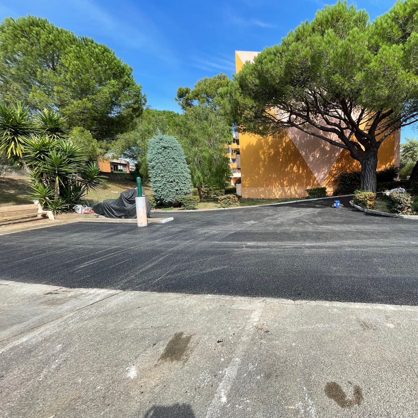 Parking asphalté fraîchement goudronné devant un immeuble d'appartements jaune et des arbres, par une journée ensoleillée.