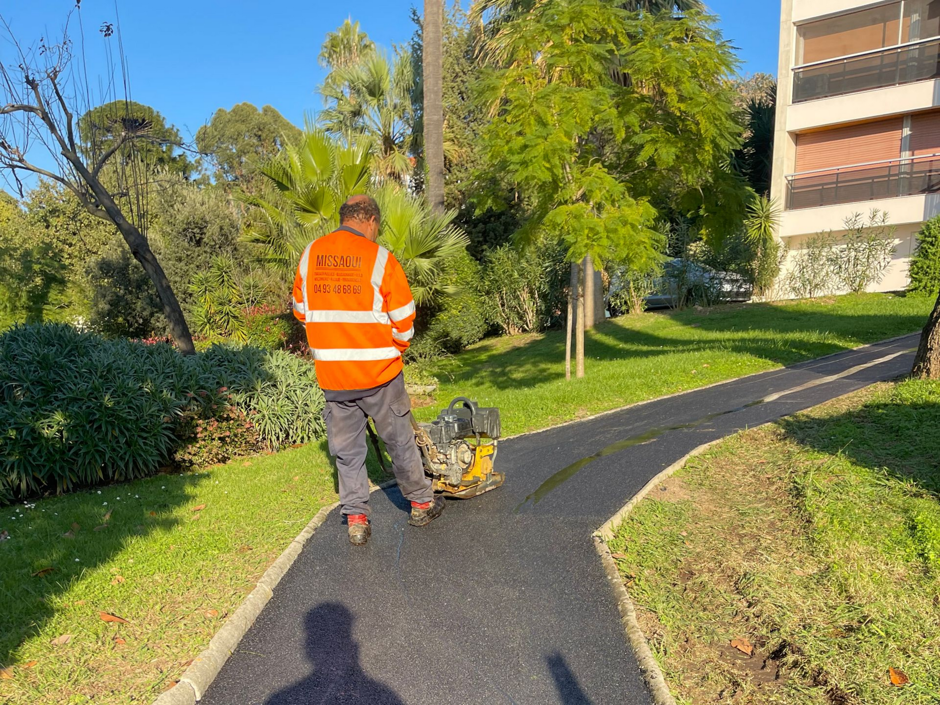 Dans un parc, une personne vêtue d'un gilet orange compacte l'asphalte d'une allée à l'aide d'une plaque vibrante.