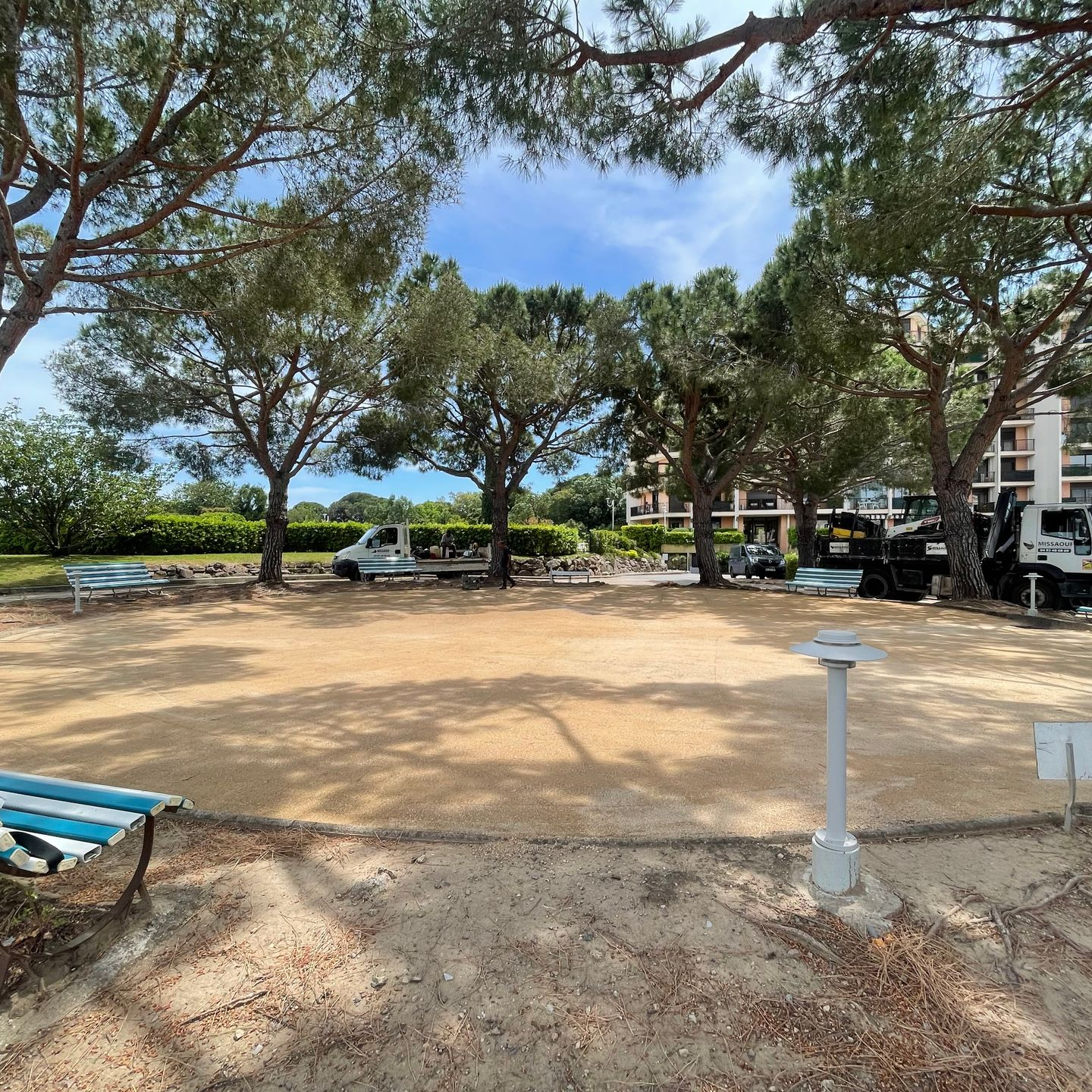 Terrain de pétanque sablonneux entouré d'arbres, de bancs et de véhicules stationnés sous un ciel bleu.