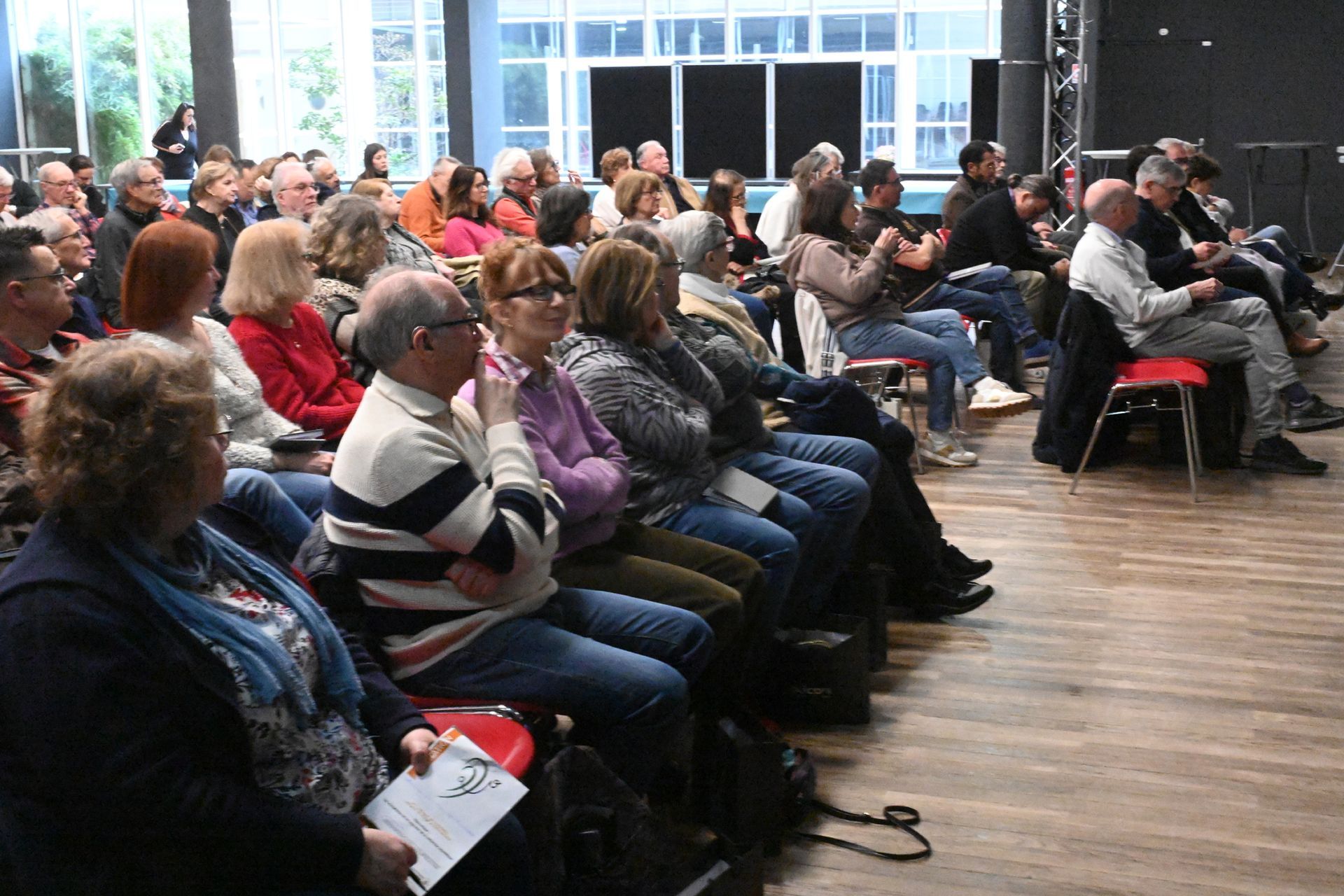 Public assis dans une salle, regardant une présentation près de grandes fenêtres