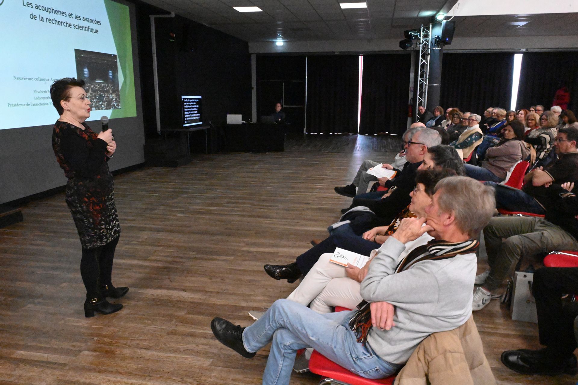 Un présentateur s’adresse à un public assis dans une salle de conférence, avec une diapositive projetée sur la gauche