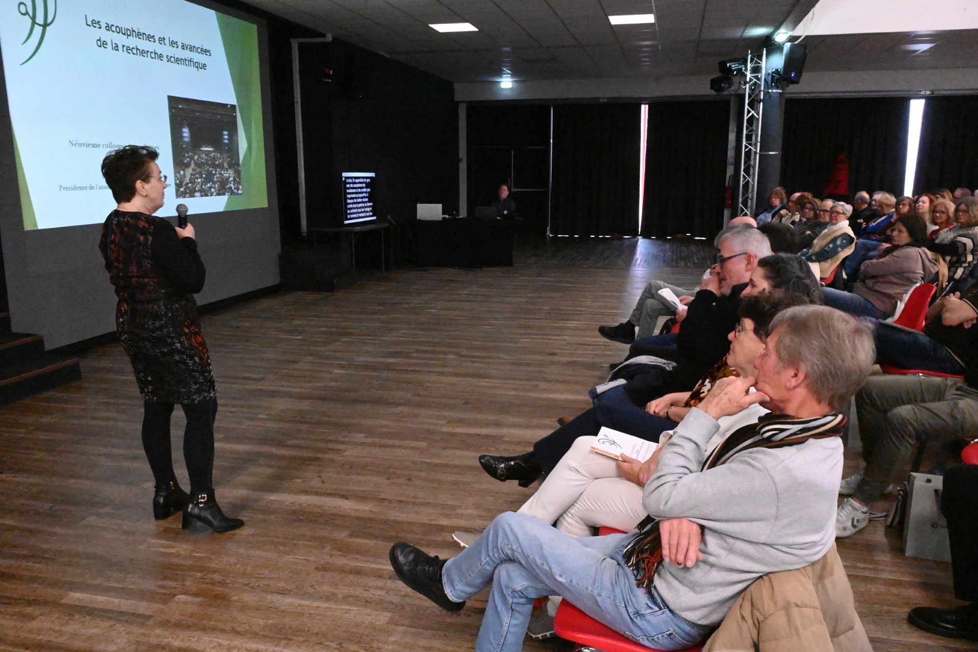 Un présentateur s’adresse à un public assis dans un auditorium sombre pendant une conférence ou une présentation