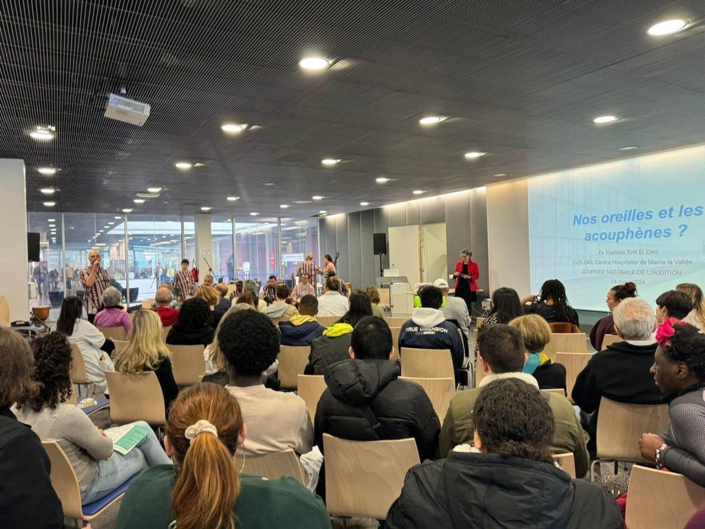 Public lors d'une présentation. Une femme prend la parole devant un écran. Plusieurs personnes sont assises ou debout dans la salle.