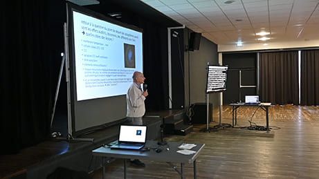 Un homme fait une présentation, debout près d'un écran affichant du texte et des images. Espace réservé au public dans une grande salle.