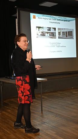 Une femme fait une présentation, debout près d'une table, regardant le public, pointant du doigt un écran de projection.