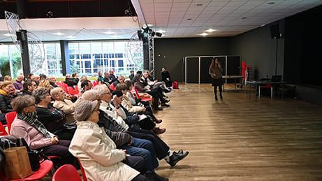 Le public est assis et écoute un présentateur sur scène dans une grande salle au parquet.