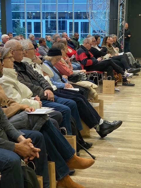 Dans une grande salle, un public est assis et écoute un orateur. Beaucoup prennent des notes.