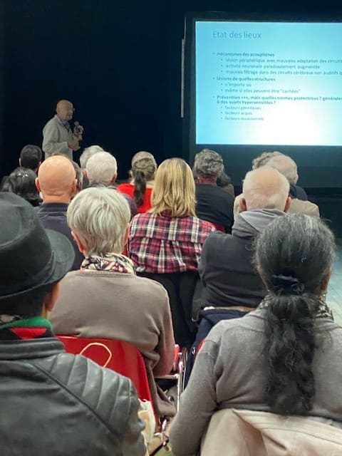 Dans un amphithéâtre, le public écoute une présentation sur un écran. Un homme parle dans un microphone.