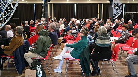 Public assis sur des chaises rouges lors d'un événement, face au public. Fond sombre, éclairage de scène.