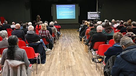 Le public, assis sur des chaises rouges, écoute une présentation sur écran dans une salle obscure.