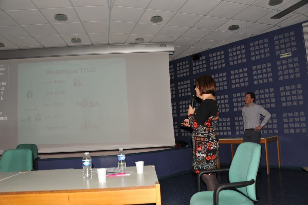 Une femme prend la parole lors d'une conférence. Elle tient un microphone, un écran de projection se trouve derrière elle. Un homme se tient à ses côtés.