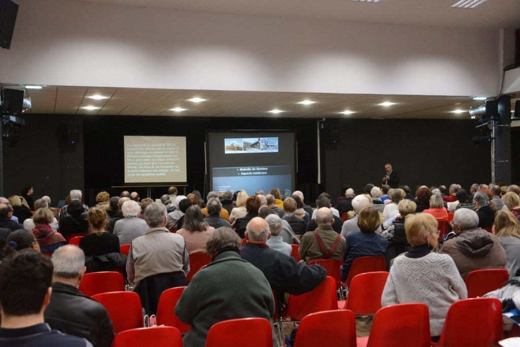 Public assis sur des chaises rouges lors d'une présentation, orateur sur scène, deux écrans affichant du texte et des images.