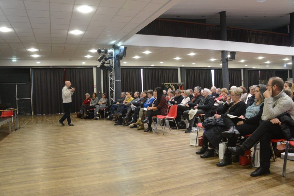 Un homme s'adresse à un public assis dans une grande salle avec un plancher et une scène en bois.