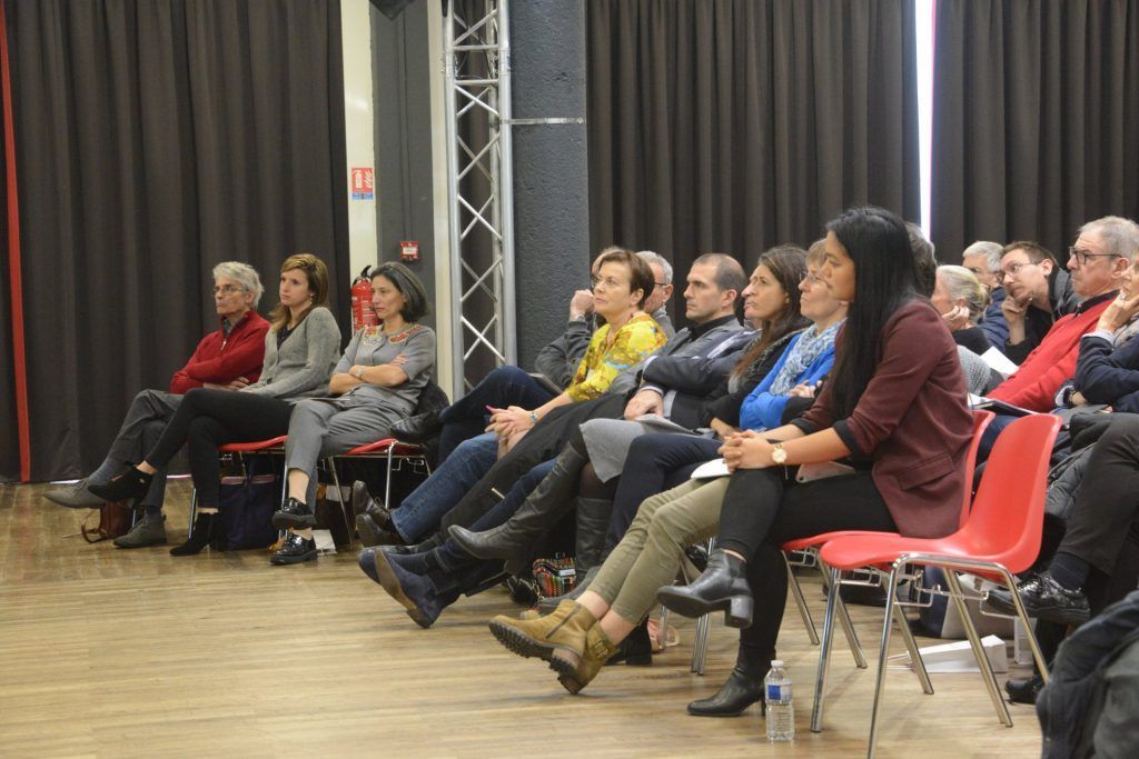 Le public, assis sur des chaises rouges, écoute attentivement dans une grande salle.