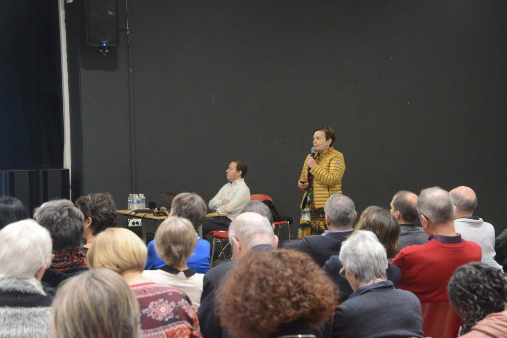 Une personne prend la parole devant un auditoire dans un amphithéâtre. Une autre personne est assise à proximité. Fond sombre.