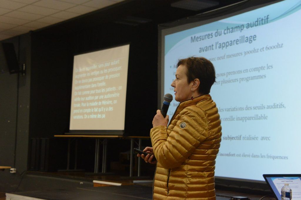 Une personne prend la parole lors d'une présentation, deux diapositives sont projetées. Veste jaune, microphone à la main.