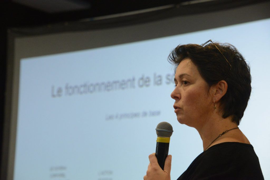 Une femme prend la parole lors d'une présentation, micro en main. Une diapositive en arrière-plan affiche « Le fonctionnement de la... »