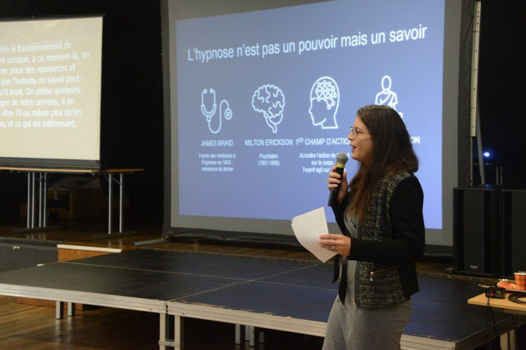 Une femme présente sur scène, micro en main, avec un écran derrière elle affichant des schémas et du texte.