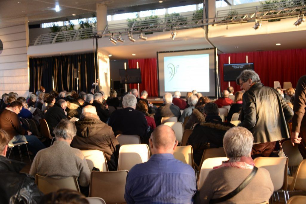 Public assis dans une salle de conférence, regardant une présentation sur un écran. Rideaux rouges, hauts plafonds.