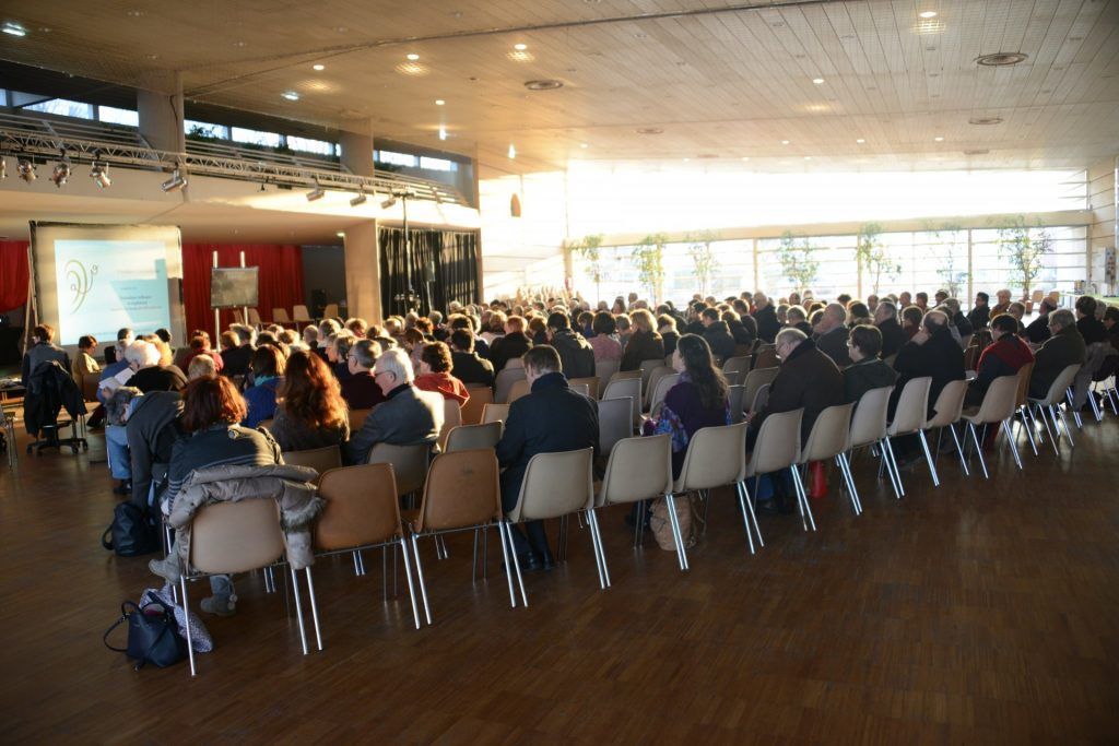 Grande salle de réception où des personnes sont assises face à un écran de projection. La lumière du soleil y pénètre abondamment.