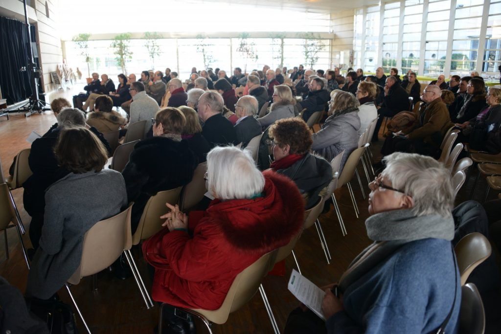 Dans une grande salle vitrée, un public assiste à un événement. Les chaises sont occupées.
