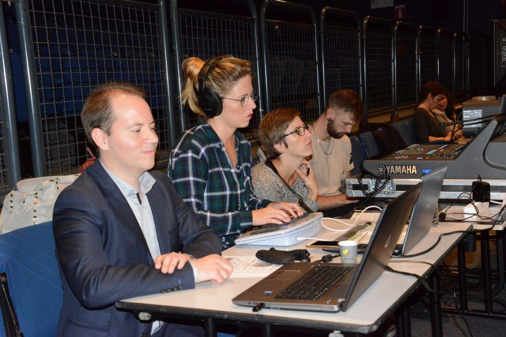 Des personnes assises à une table travaillent avec des ordinateurs portables, des claviers et une table de mixage, probablement lors d'un événement en direct.