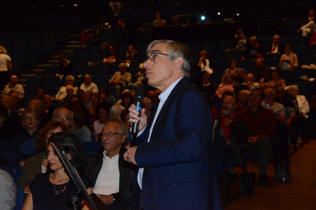 Un homme en costume parle dans un microphone, s'adressant à un public assis dans un auditorium plongé dans l'obscurité.
