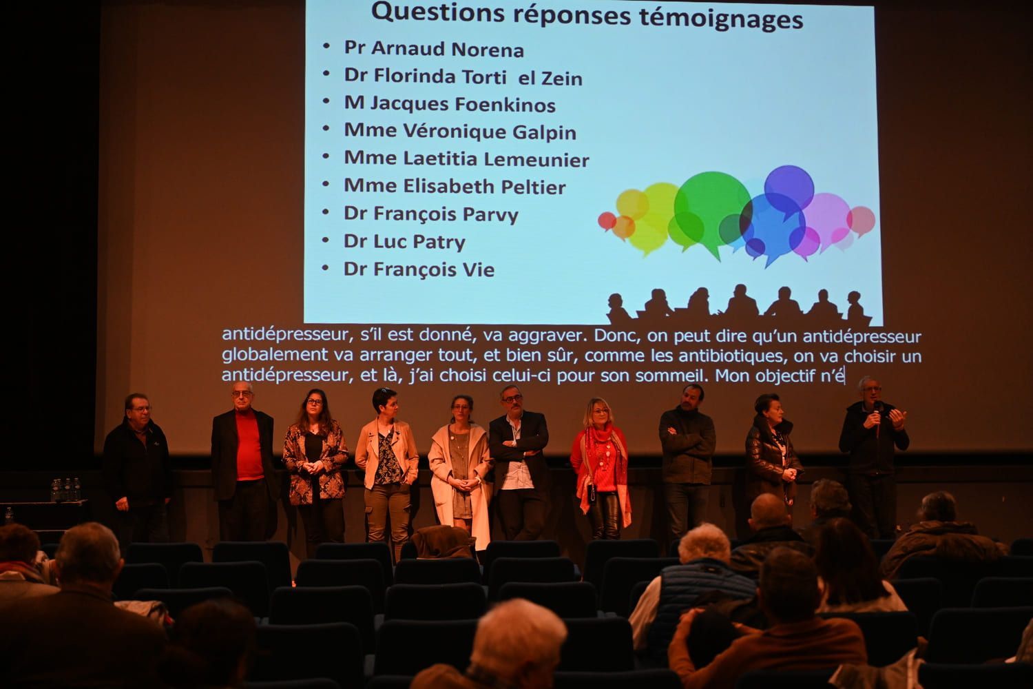 Table ronde sur scène, avec projection sur écran de la liste des intervenants. Public assis dans un théâtre.