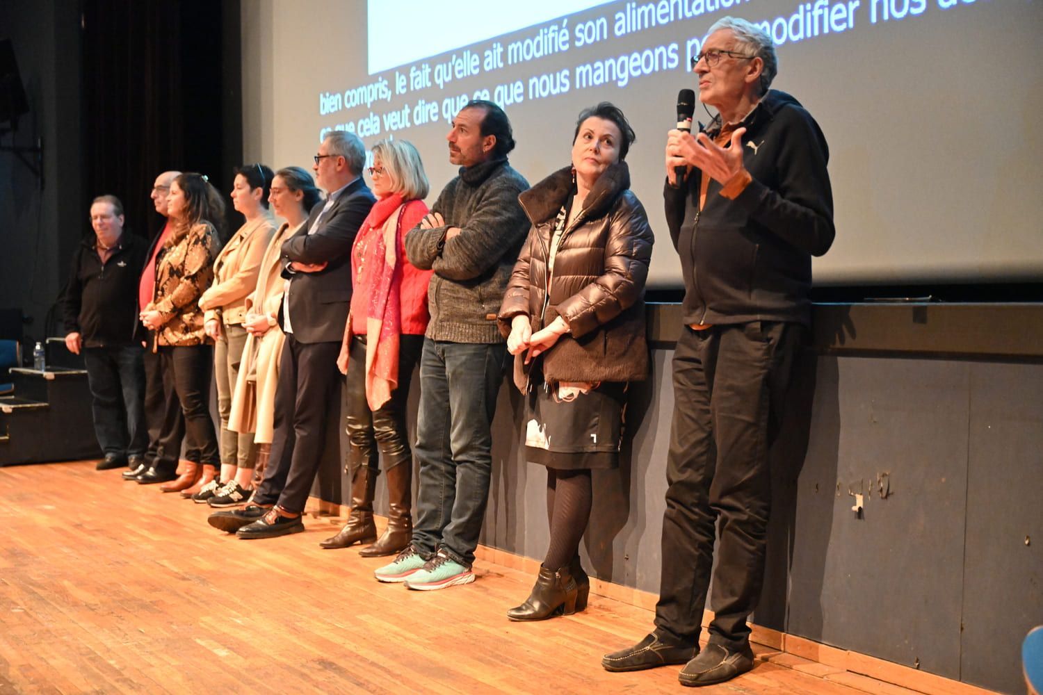 Un groupe de personnes se tient sur scène ; certaines écoutent, une autre parle dans un microphone. Fond sombre avec du texte.