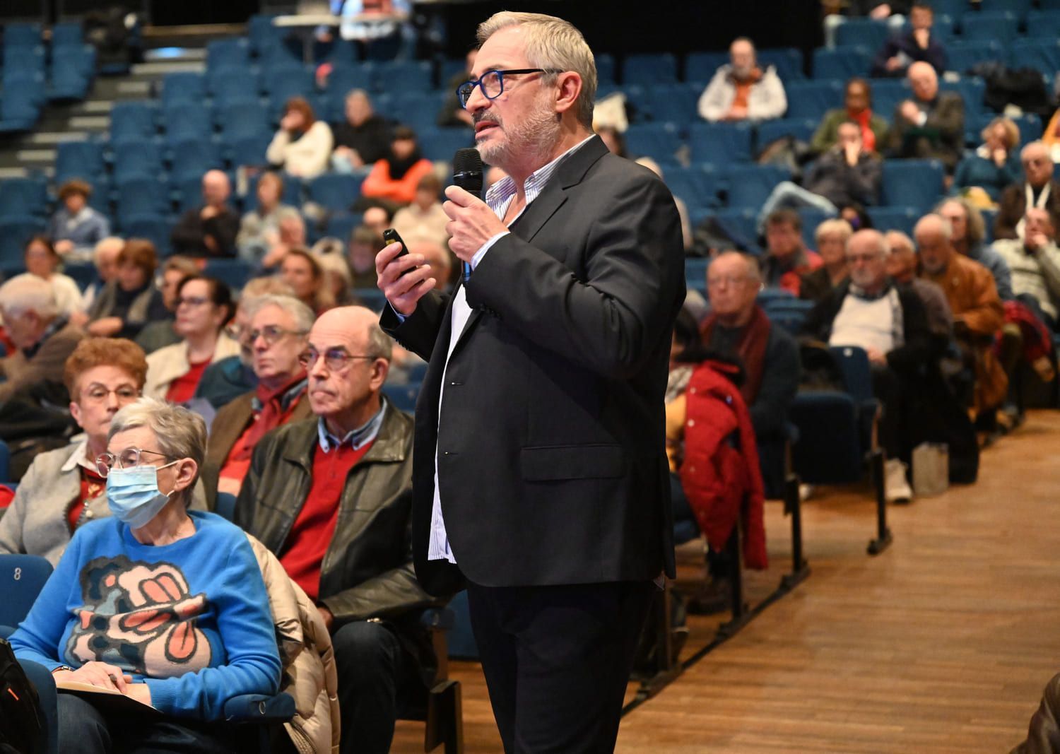 Un homme parle au micro devant un public dans un grand auditorium aux sièges bleus.
