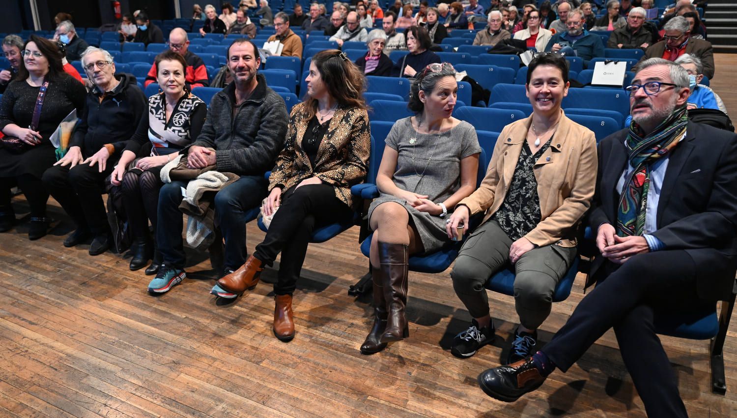 Des personnes assises dans un auditorium écoutent. Certaines regardent la caméra. Lambris et chaises bleues.
