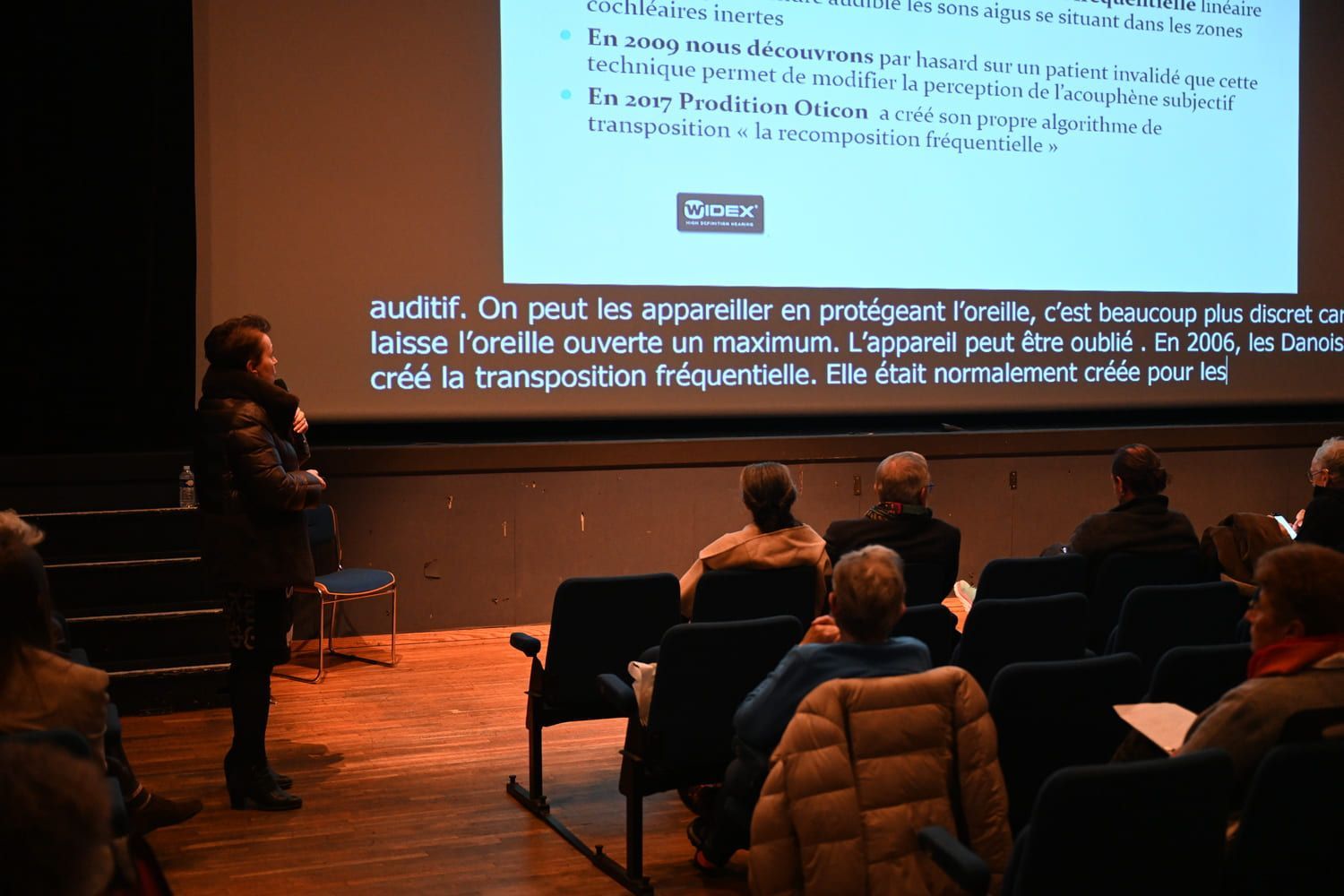 Une personne prend la parole devant un écran affichant du texte. Les membres du public sont assis sur des chaises, face à l'orateur.