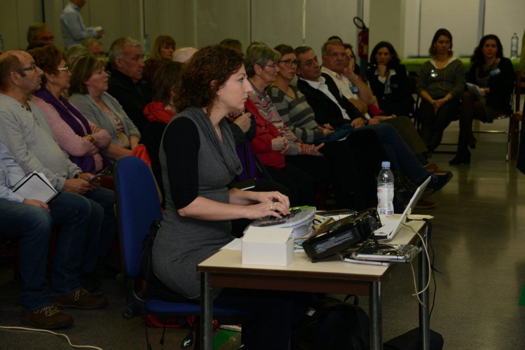 Une femme tape à un ordinateur portable, présentant un exposé à un public assis dans une pièce baignée de lumière.
