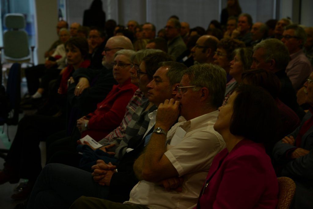Public assis et écoutant dans un amphithéâtre. Des personnes vêtues de tenues variées, certaines levant les yeux au ciel.