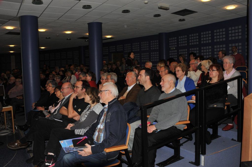 Le public est assis dans un auditorium aux murs bleus, écoutant une présentation.