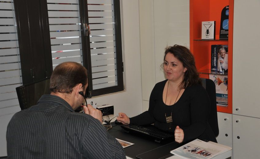 Une femme discute avec une personne assise à un bureau. Bureau noir, mur orange, décor neutre.