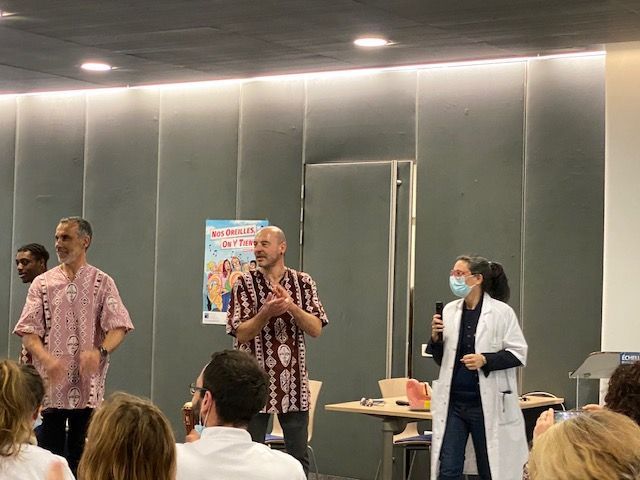 Trois hommes et une femme sont sur scène. Un homme applaudit, un autre sourit. La femme parle dans un micro. Une affiche est visible.