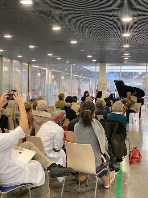 Des spectateurs assis écoutent un pianiste et un violoniste. Un concert en plein air, à la lumière naturelle.
