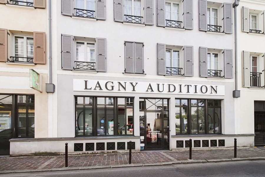 Façade du magasin Lagny Audition avec volets gris et enseigne sur un bâtiment blanc, vue de la rue.