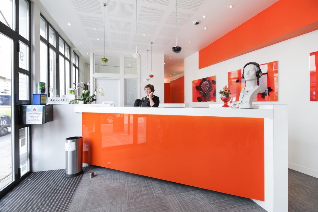 Bureau d'accueil avec façade orange, comptoir blanc et une personne derrière. Œuvre d'art au mur.