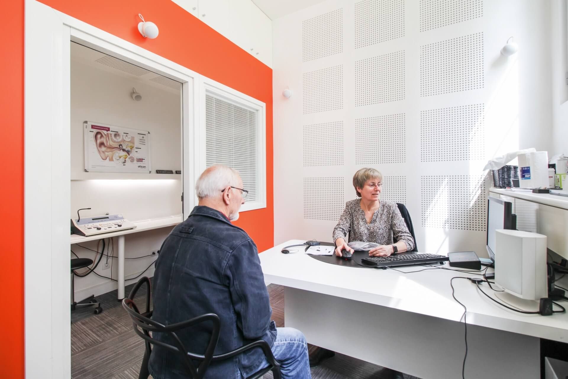 Un homme passe un test auditif avec un audioprothésiste dans une cabine insonorisée orange et blanche.