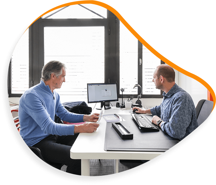 Deux hommes assis à un bureau dans un bureau, l'un regardant des documents, l'autre à un ordinateur, tous deux en chemise bleue.