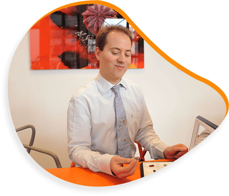 Homme souriant, assis à une table orange, chemise bleu clair, cravate à motifs. Œuvre d'art moderne au mur en arrière-plan.