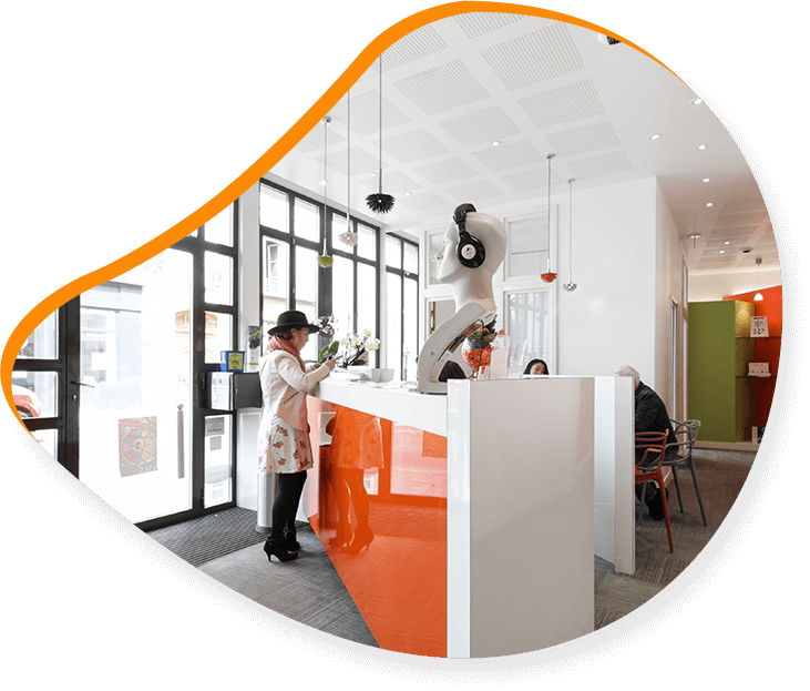 Un comptoir d'accueil moderne blanc et orange où deux personnes interagissent. Un espace intérieur lumineux.