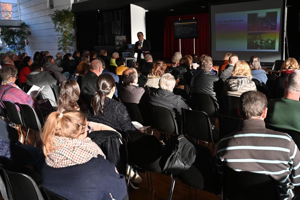 Le public, assis, écoute une présentation faite par un homme à un pupitre, des diapositives étant projetées sur un écran.