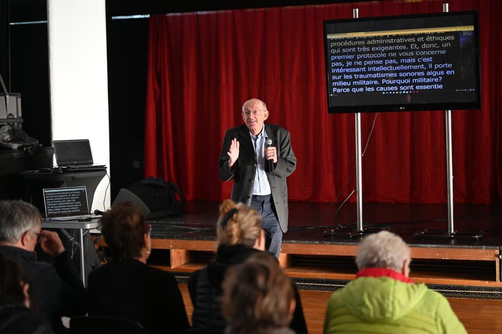 Un homme prend la parole lors d'une présentation, gesticulant, avec un écran affichant du texte derrière lui ; le public est assis devant lui.